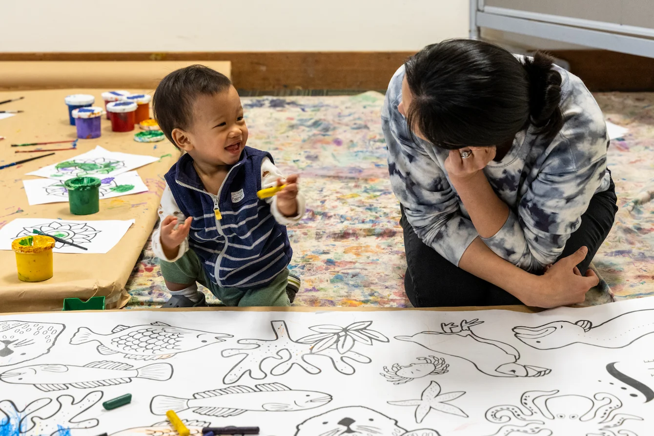 A caretaker and a young child sit in front of a large coloring sheet art project during an activity at the Seattle Aquarium.