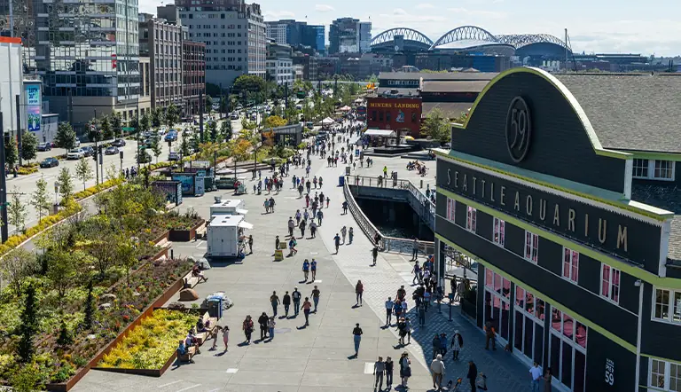 Seattle's Waterfront Park, as seen from atop the Overlook Walk looking south. The Seattle Aquarium's Pier 59 building is featured to the right, with pathways, bike paths, and green spaces along the park.